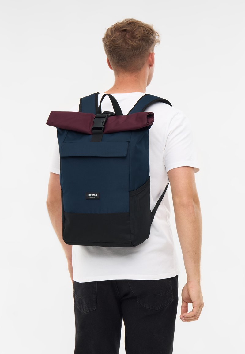 Tengerészek hátizsákja bordó, felgombolható felső résszel �és fekete aljjal. Állítható pántokkal és elülső zsebbel rendelkezik, tartós anyagból készült.