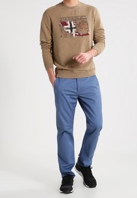Felpa marrone con una toppa grafica che presenta un design rosso e nero, abbinata a pantaloni blu e sneakers nere.