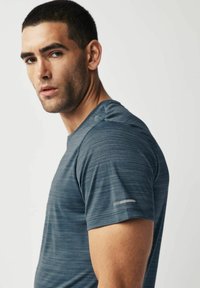 Camiseta deportiva gris-azul con un tejido texturizado que absorbe la humedad. Presenta mangas cortas y un acento reflectante en el hombro izquierdo.