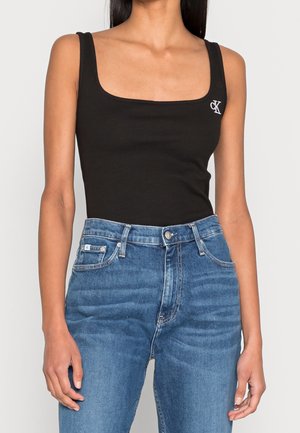 Woman wearing a black sleeveless square-neck top with white logo near shoulder, paired with high-waisted blue jeans.