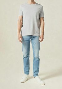 Mann trägt ein hellgraues kurzärmeliges Hemd, hellblaue Jeans und weiße Sneaker, steht vor einem einfarbigen beigen Hintergrund.