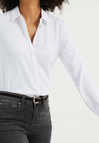Bílá košile na knoflíky s límečkem, vyrobená z lehkého materiálu; spárovaná s tmavě šedými džínami a černým opaskem se zlatou sponou.