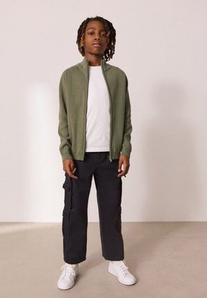 Mladá osoba so stredne dlhými dredmi, oblečená v olivovo zelenom svetri na zips, bielom tričku, čiernych cargo nohaviciach a v bielych teniskách.