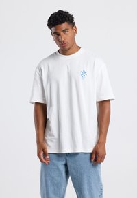 White cotton t-shirt with short sleeves and round neckline. Features a small blue embroidered design on the left chest. Casual fit.