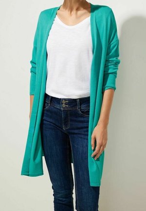 Femme portant un cardigan ouvert couleur sarcelle, un t-shirt blanc à col rond et un jean bleu foncé, debout devant un fond clair uni.