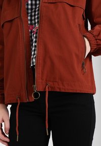 Chaqueta utilitaria de color óxido con cremallera, puños elásticos y dobladillo con cordón ajustable. Cuenta con dos bolsillos frontales y un forro interior de cuadros.