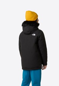 Μαύρο μπουφάν puffer με κουκούλα επενδεδυμένη με γούνα, που έχει λευκό λογότυπο στον αριστερό ώμο, συνδυασμένο με μπλε παντελόνι και κίτρινο σκουφάκι.