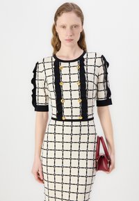 Rochie ajustată de culoare crem cu un model în carouri negre, nasturi aurii și accente negre. Modelul ține o geantă mică roșie. Material texturat.