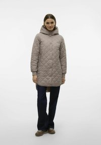 Gekwiet, neutrale beige jas met een capuchon, drukknoopsluiting en lange mouwen; gecombineerd met donkerblauwe, uitlopende jeans en bruine schoenen.