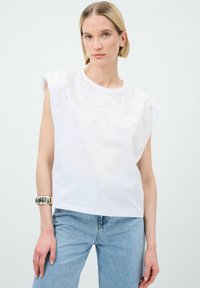 Vit kortärmad topp med upphöjda dekorativa pärlor, rund halsringning och avslappnad passform, i kombination med ljusblå jeans. Slät bomullstextur.