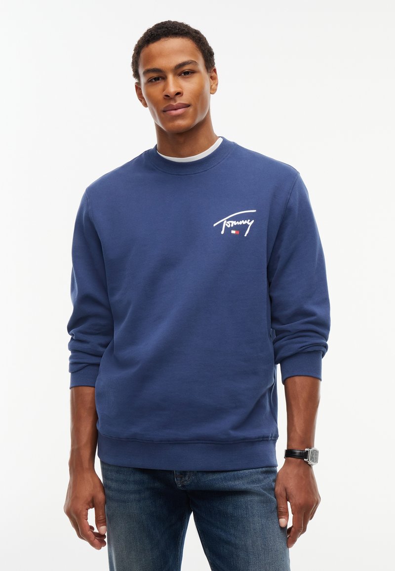 Homme portant un sweat-shirt bleu à manches longues avec le logo "Tommy" sur la poitrine et un jean bleu, debout devant un fond blanc uni.