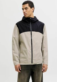 Veste légère en tissu synthétique présentant un design à blocs de couleur en noir et beige, avec une fermeture éclair à l'avant et une capuche ajustable.