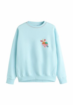 Frau trägt ein hellblaues Sweatshirt mit einer bunten Eisschüssel und dem auf der Brust eingestickten Schriftzug „Gelato please“.