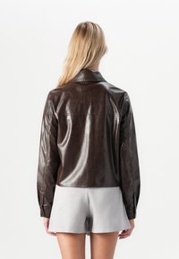 Brun læderjakke med en glat overflade, der har en klassisk krave, lange ærmer og en kortet kant, parret med grå shorts.