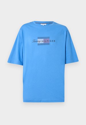 Személy kék Tommy Hilfiger pólóban, világoskék farmerben, bézs sportcipőben és kék sapkában, egyszerű fehér háttér előtt áll.