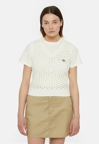 Gilet bianco a maglia con scollatura a V e design a tramatura aperta, indossato sopra una t-shirt bianca, abbinato a una gonna beige. Caratterizzato da un piccolo logo.