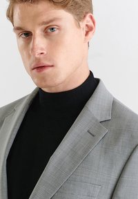 Szary w kratę marynarka z klapami typu notch, zestawiona z czarnym golfem. Gładka tekstura, delikatne przeszycia i dopasowany krój.