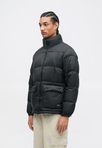 Μαύρο μπουφάν με ψηλό γιακά, με ραφές, δύο μπροστινές τσέπες και ελαστικές μανσέτες, φτιαγμένο από ελαφρύ, αδιάβροχο ύφασμα.