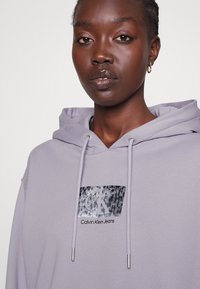 Jasnofioletowa bluza z kapturem z regulowanym sznurkiem, z teksturowaną naszywką logo CK i napisem "Calvin Klein Jeans" w kolorze czarnym. Wykonana z bawełny, o luźnym kroju.