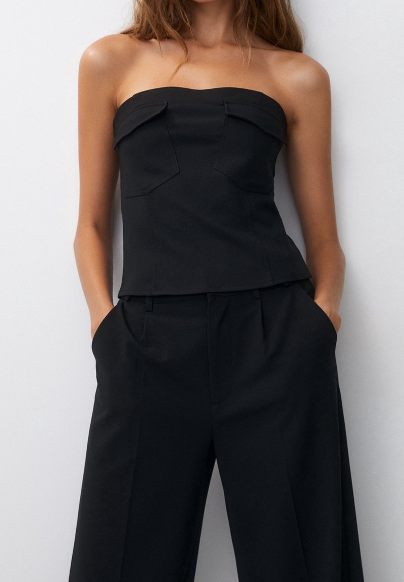 Femme portant un haut noir sans bretelles avec deux rabats à l'avant et un pantalon noir large taille haute, les mains dans les poches.
