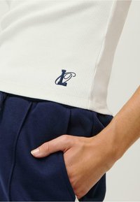 Bijela rebrasta majica bez rukava s mornarskom izvezenom logotipom na rubu, u kombinaciji s mornarskim kratkim hlačama od tkanine. Close-up prikazuje detalj ruke u džepu.