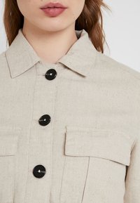 Beżowa lniana koszula z guzikami, z kolnierzykiem, dwoma przednimi kieszeniami i czarnymi guzikami. Tekstura wydaje się subtelna i lekko szorstka.