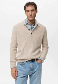 Beżowy sweter z kołnierzykiem, trzema guzikami i teksturowaną tkaniną, noszony na jasnoniebieskiej koszuli i zestawiony z jasnoniebieskimi dżinsami.
