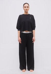 Γυναίκα που στέκεται ξυπόλητη, φορά χαλαρό μαύρο παντελόνι και κοντό μακρυμάνικο μαύρο τοπ, με εμφανή μέση Polo Ralph Lauren.