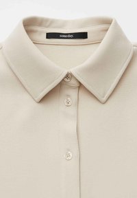 Polo beige con collo a risvolto, caratterizzata da una patta con tre bottoni bianchi e una texture del tessuto morbida e liscia.