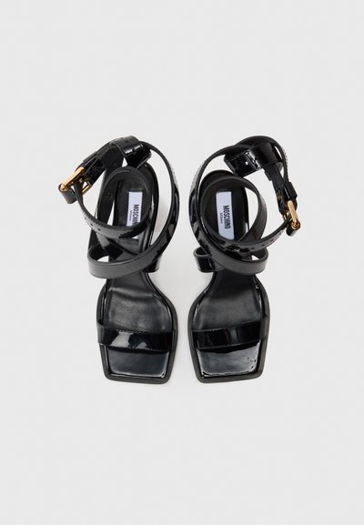 Sandale cu toc înalt din piele patentă neagră, cu vârf pătrat, curea frontală unică și curele pentru gleznă ajustabile cu catarame aurii, etichetate Moschino.