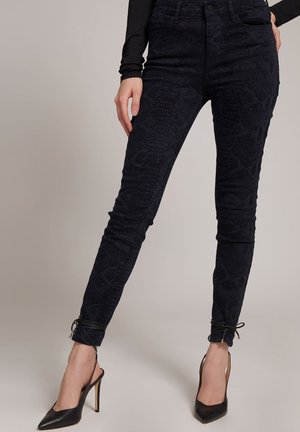 Jeans ajustados de un azul oscuro con una textura de estampado de serpiente, que presentan una cintura de altura media, bolsillos frontales y un detalle atado en el tobillo.
