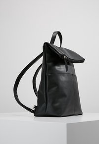 Černý kožený batoh s horním zipem na klopě, nastavitelnými ramenními popruhy a malým držadlem, postavený vzpřímeně na bílém povrchu.