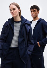 Γυναίκα και άνδρας που φορούν μπλε σκούρα φούτερ με κουκούλα και ριγέ πουκάμισα, στέκονται μπροστά σε ένα απλό ανοιχτόχρωμο φόντο, και οι δύο με τα χέρια στις τσέπες.