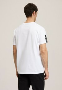 Biała bawełniana koszulka z krótkim rękawem, z czarną metką z tkaniny na lewym rękawie. Prosty design, luźny krój, gładka faktura.