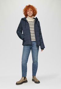 Marineblauwe pufferjack met capuchon, ritssluiting aan de voorkant en elastische manchetten, gecombineerd met een gestreepte trui en blauwe jeans, afgewerkt met bruine enkellaarzen.