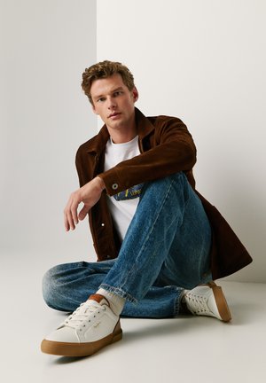Jeune homme aux cheveux bouclés assis en tailleur, portant des baskets blanches, un jean bleu, un t-shirt blanc et une veste marron sur un fond uni.