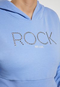 Felpa azzurra chiara con tasca a marsupio. Caratterizzata da "ROCK" in lettere argentate con borchie e "my soul" in bianco, realizzata in tessuto morbido e testurizzato.