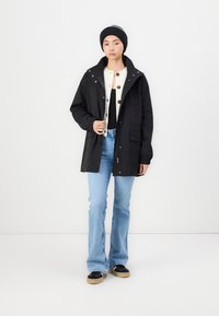 Parka noir imperméable avec capuche, dotée d'un devant boutonné, de deux poches latérales et d'une coupe décontractée, associée à un jean évasé bleu clair et des chaussures noires.