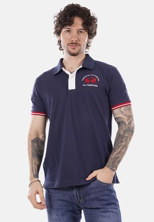 Uomo con capelli ricci che indossa una polo blu navy con logo rosso e bianco, polsini delle maniche a righe rosse e bianche, e jeans blu, in piedi con una mano in tasca.