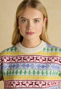 Strickpullover mit weißem Grund und bunten Mustern in Rot, Grün, Blau und Gelb, darunter Rehe und Bäume, sowie einem gerippten Kragen.