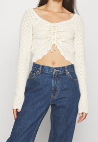 Top court en crochet crème avec un décolleté carré, un devant à cordon et un ourlet en forme de vagues. Associé à un jean bleu taille haute.