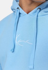 Sudadera azul claro hecha de una tela suave, con un logo bordado en blanco en el pecho y cordones con ojales negros.