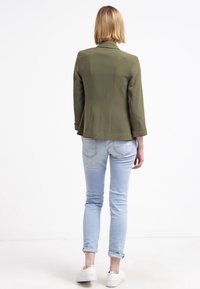 Olivově zelený blazer s přizpůsobeným střihem, mírně strukturovanými rameny a ohrnutými manžetami. V kombinaci se světle modrými džíny a bílými teniskami.
