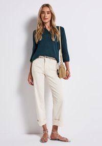 Femme portant un haut à manches longues bleu sarcelle, un pantalon crème taille haute avec des revers, portant un sac à bandoulière beige et des sandales blanches à lanières.