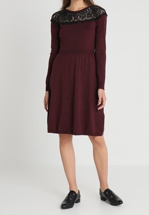 Vrouw draagt een knielange bordeauxrode jurk met lange mouwen en zwarte kantdetails bij de hals, gecombineerd met zwarte instapschoenen.