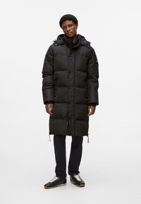 Abrigo de plumas negro con capucha, con textura acolchada, botones al frente y cordones ajustables en el dobladillo. Llevado sobre pantalones oscuros y zapatos.