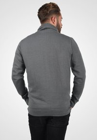 Sudadera gris con un cuello alto, mangas largas y puños acanalados, que presenta una tela texturizada y un ajuste relajado, combinada con jeans oscuros.