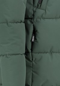 Dunkelgrüne Steppjacke mit einem quilted Design, ausgestattet mit einer Reißverschlusstasche und einem Etikett am Ärmel mit der Aufschrift "SBLVI." Weicher, strukturierter Stoff.