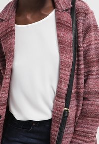 Detailní záběr osoby, která má na sobě bílý top, vínový texturovaný cardigan, tmavé kalhoty a černý popruh přes rameno k kabelce.