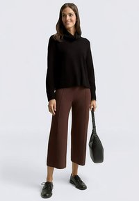 Black ribbed sweater with a cowl neck, brown wide-leg pants, and black lace-up shoes. A black shoulder bag is held in one hand.